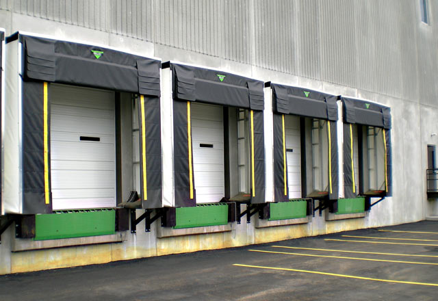 Four loading dock doors with black canopies on a warehouse, next to an empty parking area with yellow lines.