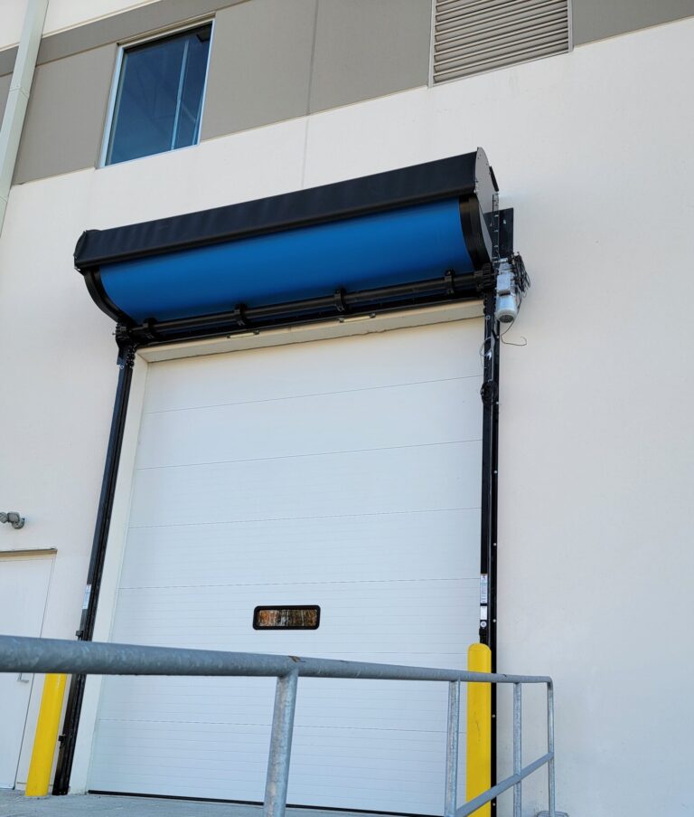 A white industrial loading dock door with a blue awning and yellow protective posts on each side.