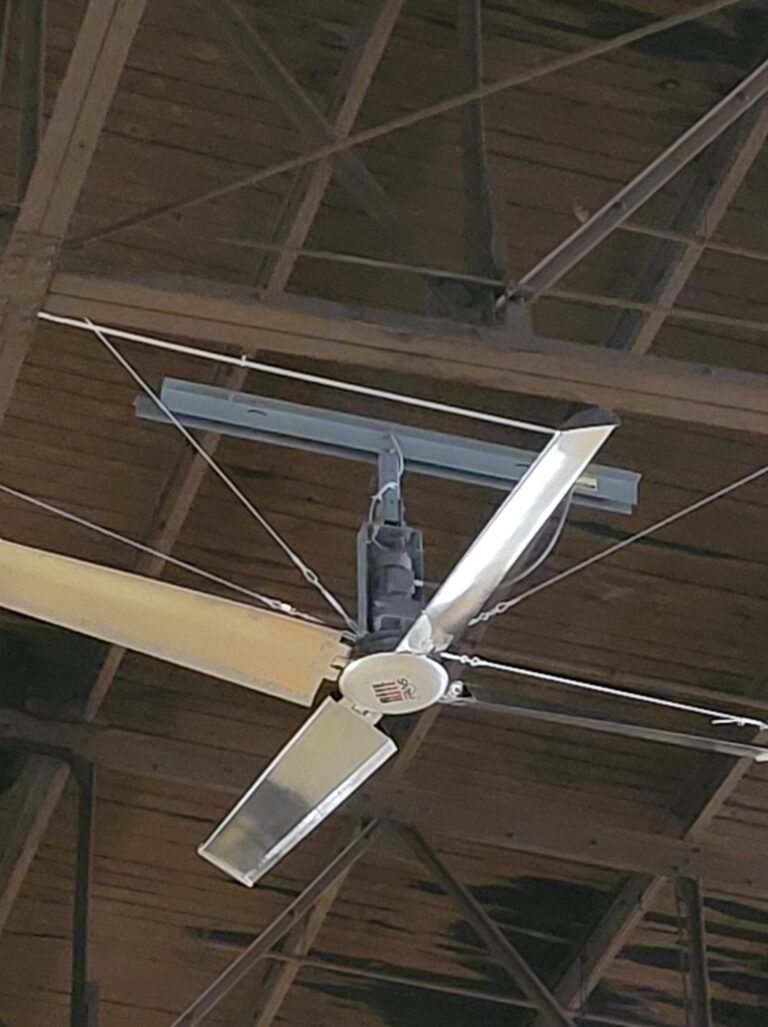 Large industrial ceiling fan with metal blades mounted on a wooden ceiling with exposed beams.