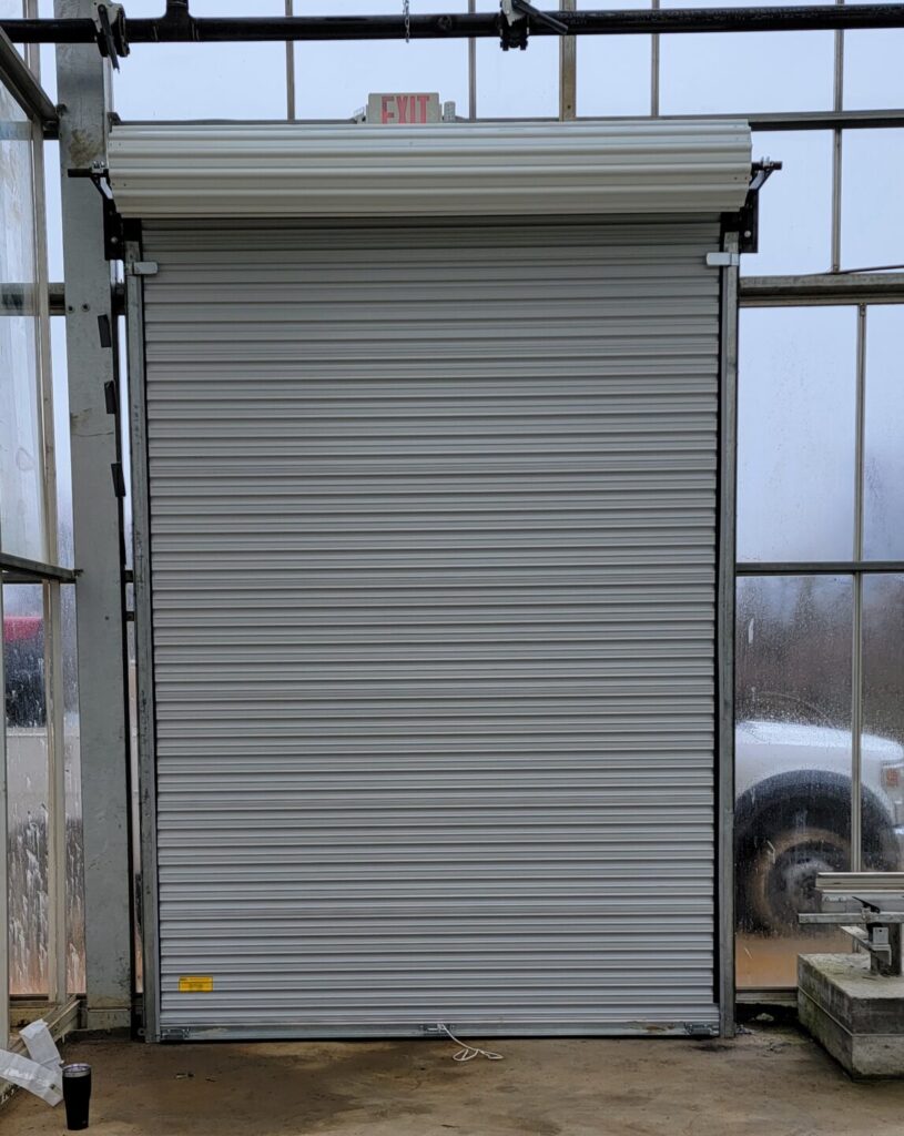 A closed metal roll-up door inside a glass building, with an exit sign above it and a vehicle partially visible outside.