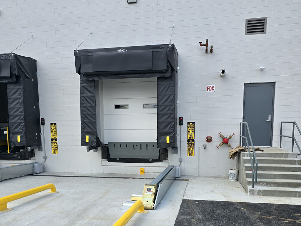 A loading dock with a closed overhead door, safety bumpers, and a gray door with steps to the right.