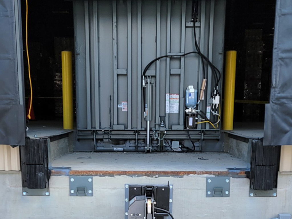 A dock leveler at a loading bay, with hydraulic equipment and yellow safety bollards on each side.