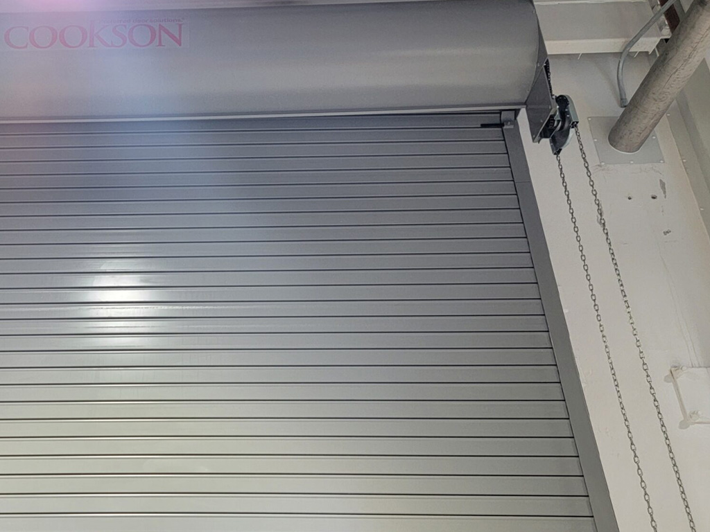 A close-up of a metallic roll-up garage door with a chain mechanism and COOKSON faintly visible at the top left.
