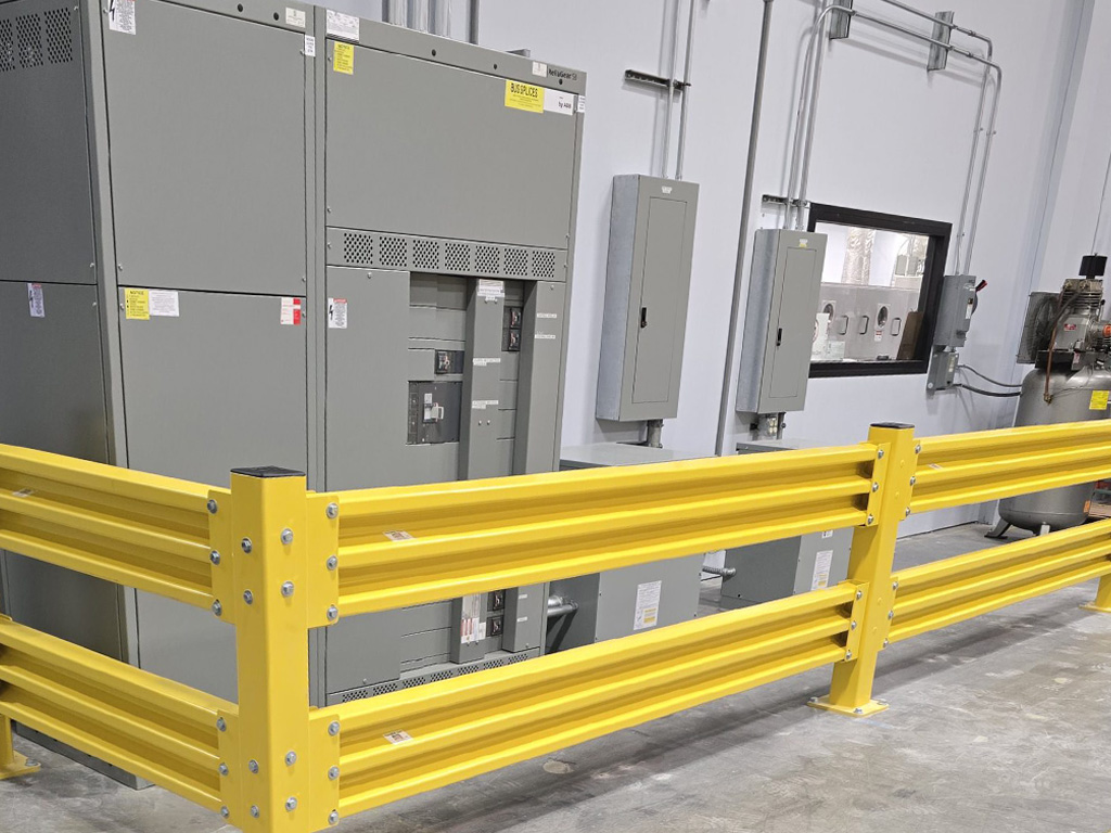 Large electrical panels behind a yellow safety barrier in an industrial facility with concrete flooring.