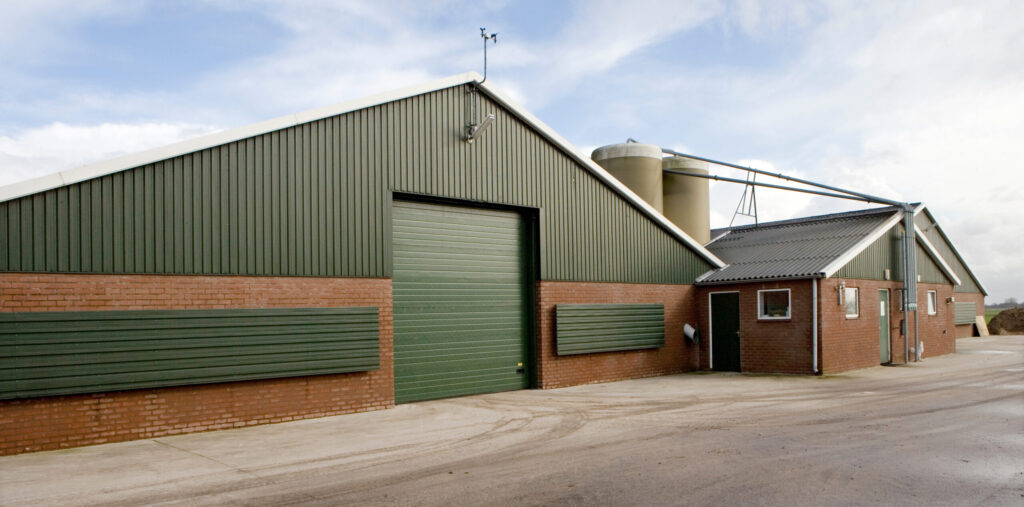 A modern agricultural warehouse with green metal siding, brick walls, and a large dock door system beside a silo.
