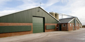 A modern agricultural warehouse with green metal siding, brick walls, and a large dock door system beside a silo.