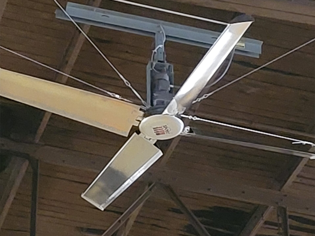 Large industrial ceiling fan with four metal blades mounted on a wooden ceiling with exposed beams.
