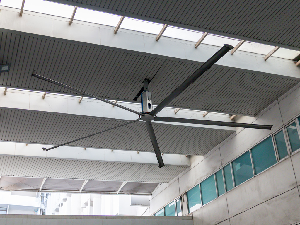 A large industrial ceiling fan with six blades is mounted on the ceiling of a modern building.