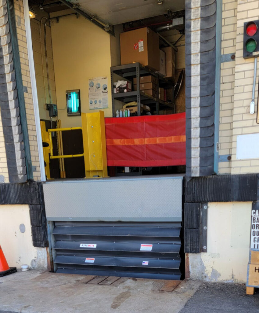 A loading dock with a closed metal barrier, shelves, and supplies visible inside, plus a red warning gate and traffic signals.