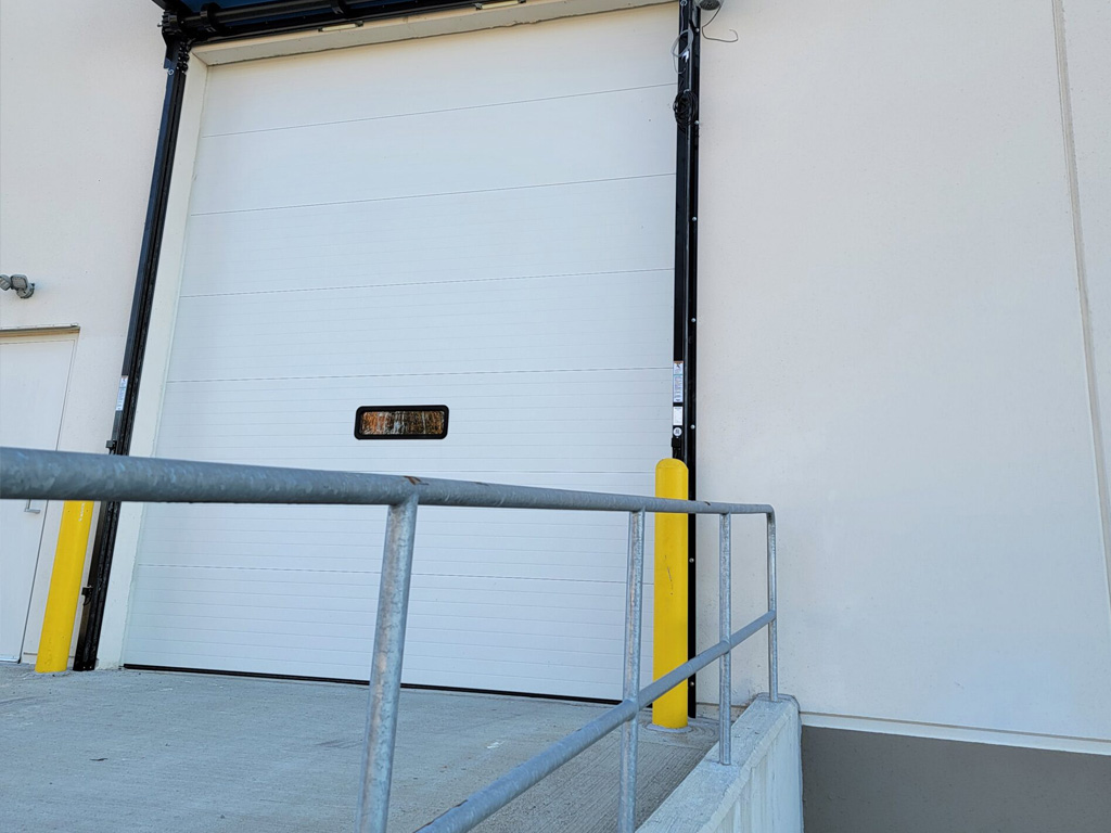 A white industrial loading dock door with a small window and yellow safety bollards on a concrete ramp.