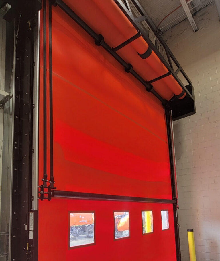 Bright red industrial roll-up door with small clear windows, installed in a warehouse with white brick walls.