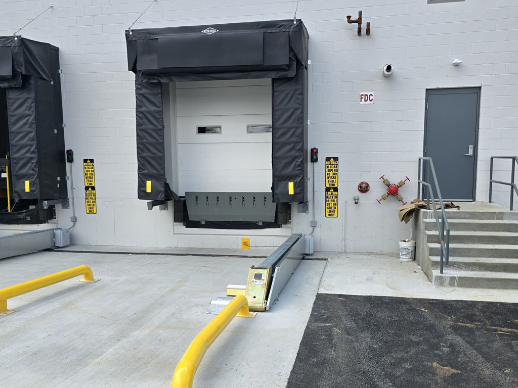 A loading dock with a closed bay door, safety barriers, warning signs, and nearby stairs with a handrail.