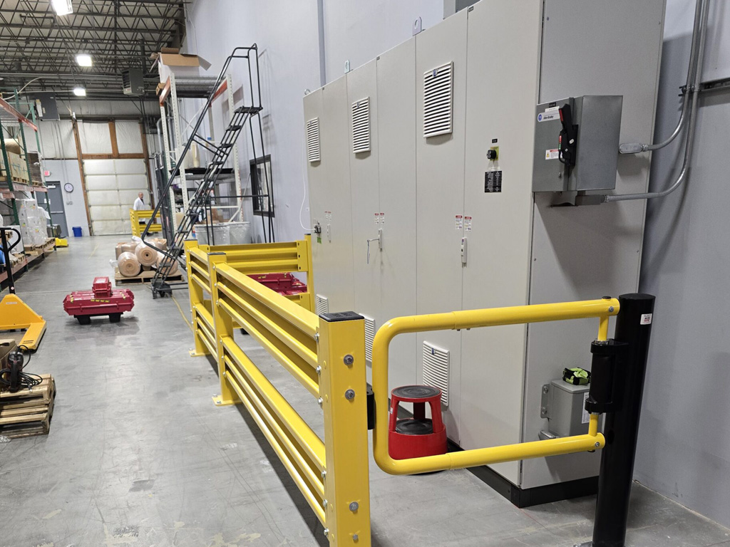 Large industrial control cabinets behind yellow safety railings in a warehouse with scattered equipment and a metal staircase.
