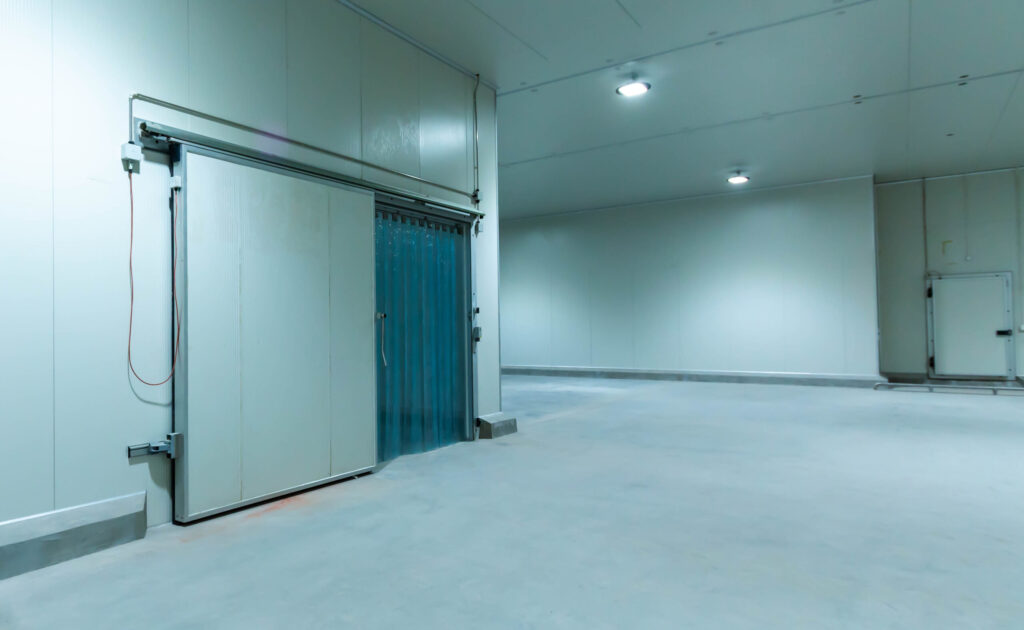 Empty industrial cold storage room with white walls, a large sliding door, and a blue strip curtain.