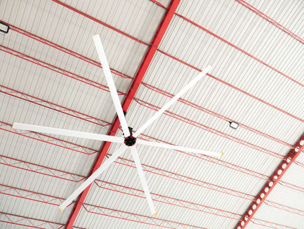 Large industrial fan installation with six white blades on a high, corrugated metal ceiling and red support beams.