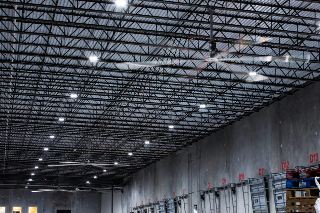 Ceiling of a warehouse with metal beams, industrial fan installation, and loading docks marked with red numbers.
