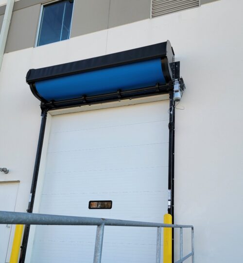 A white industrial loading dock door with a blue awning and yellow protective posts on each side.