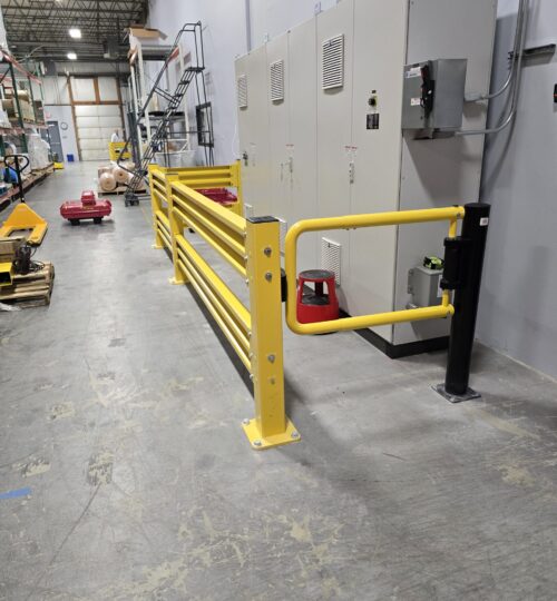Yellow safety railings are installed in a warehouse, protecting large gray electrical panels along the wall.