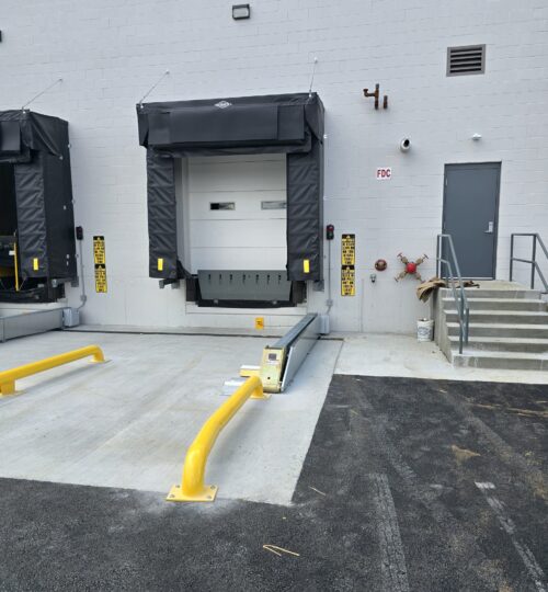 Loading dock with a damaged yellow safety barrier, steps, and a gray door on a concrete building exterior.
